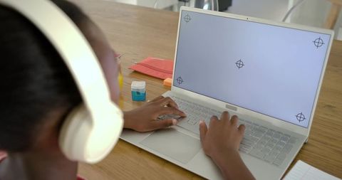 African teen studying on laptop wearing white headphones at wooden desk, typing homework