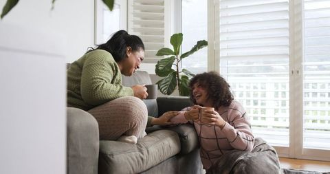 Female friends laughing and sharing coffee on cozy living room sofa