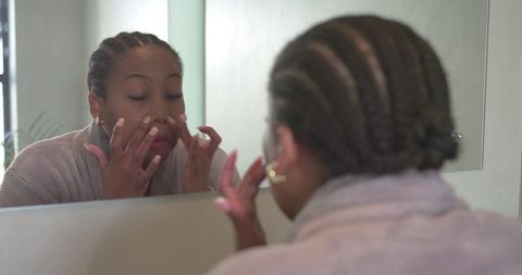 African american woman applying facial skincare at mirror wearing bathrobe and hoop earrings