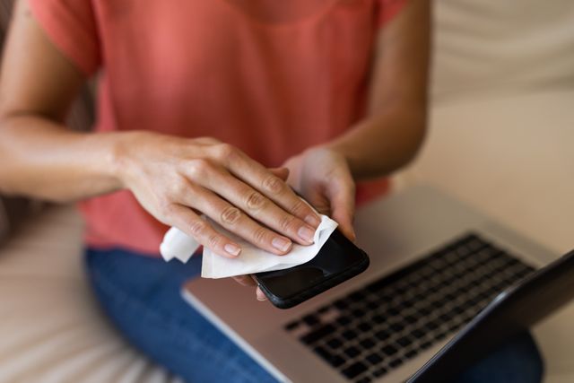 This image shows a woman cleaning her smartphone with a napkin while using a laptop. It can be used to illustrate themes of hygiene, multitasking, and technology use at home. Ideal for articles or advertisements related to cleanliness, tech maintenance, or home office setups.