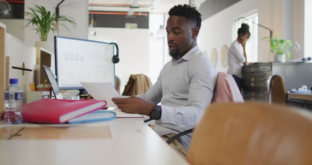 A focused African American man analyzes documents while seated at a modern office desk. This can be used for themes involving business, productivity, modern workplace, technology in business, professional analysis, and focused work.