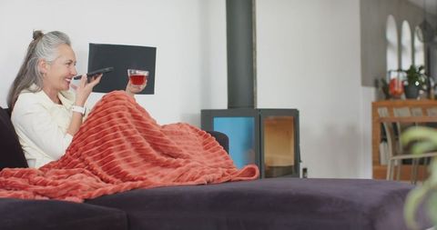 Senior woman relaxing on sofa under blanket, sipping tea and speaking on smartphone