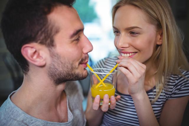 Young couple sharing a glass of orange juice with straws in a cozy cafe. They are smiling and looking at each other, creating a warm and intimate atmosphere. Perfect for use in advertisements, social media posts, or articles about relationships, dating, and lifestyle.