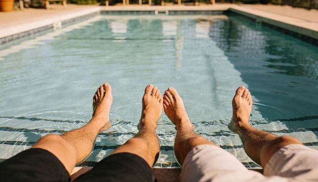 Two men relaxing at poolside dipping bare feet into sunlit water on warm summer day
