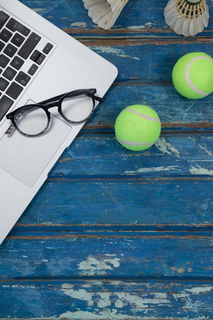 Overhead view of laptop and eyeglasses with shuttlecocks by tennis balls on blue wooden table