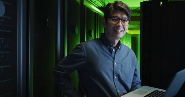 Asian IT professional working in data center surrounded by server racks and green lighting, smiling and holding a laptop. Useful for technology, engineering, data management, cyber security themes, as well as advertisements and articles about careers in IT.