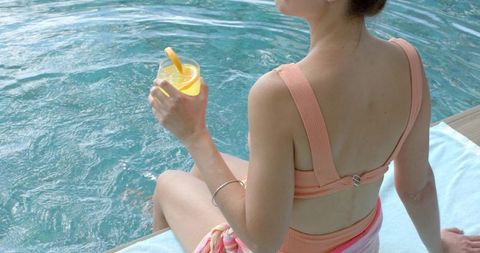 Woman sitting poolside in peach swimsuit holding citrus cocktail, summer relaxation lifestyle