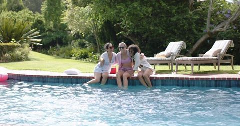 Three friends relaxing poolside dipping feet in backyard pool on sunny afternoon