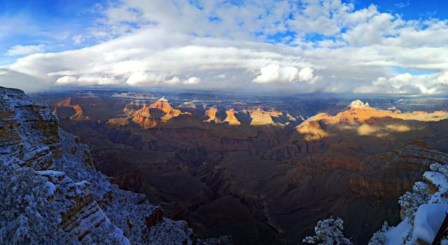 Panoramic view of the Grand Canyon at sunset during winter with snowy landscape and cloudy sky, casting shadows and highlighting the dramatic cliffs. This image is perfect for travel promotions, nature calendars, outdoor enthusiast blogs, and winter season advertisements showcasing natural marvels and adventurous destinations in Arizona.
