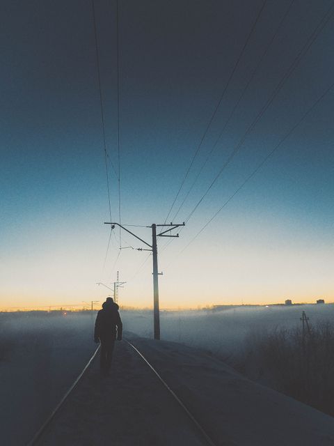 Silhouette of a person walking alone on railroad tracks in a misty urban area at dusk, surrounded by electric pylons. This image evokes feelings of solitude, mystery, and contemplation. Ideal for use in stories about journeys, exploration of the unknown, or urban solitude themes.