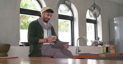 Indian man sitting on wooden kitchen counter holding mug and using tablet
