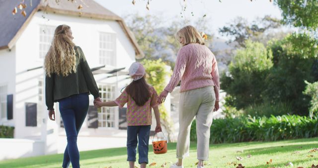Family holding hands, walking through a sunlit garden during autumn. Girl carrying Halloween pumpkin bucket, adults wearing sweaters and casual attire. Perfect for topics on family bonding, outdoor activities, autumn season, parent-child relationships, and holiday preparation.