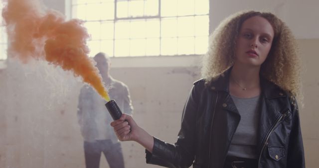 Young Caucasian woman holds a flare in an industrial setting. Her intense expression adds drama to the scene with a hazy backdrop.