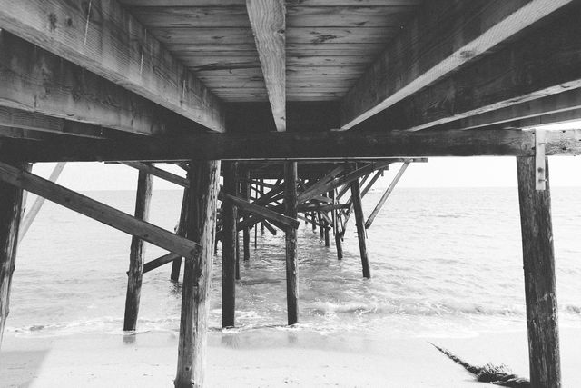 Stylized black and white image showcasing the view under a wooden pier with ocean waves hitting the sandy shoreline. Moody and nostalgic feel makes it suitable for wall art, travel blogs, coastal-themed designs, and books or magazines focused on seaside environments.