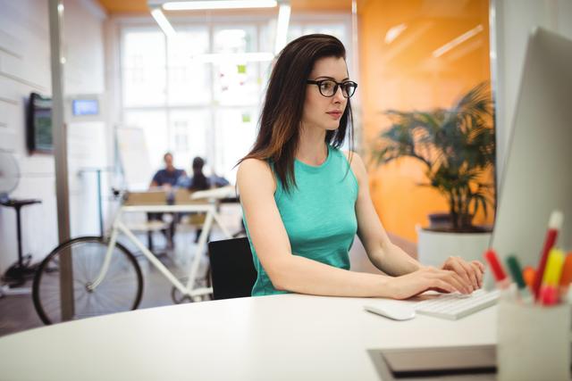 Female executive working at her desk in a modern office. She is focused on her computer screen, wearing glasses and a teal sleeveless top. Ideal for use in business, technology, and productivity-related content, showcasing a professional work environment and dedicated employee.