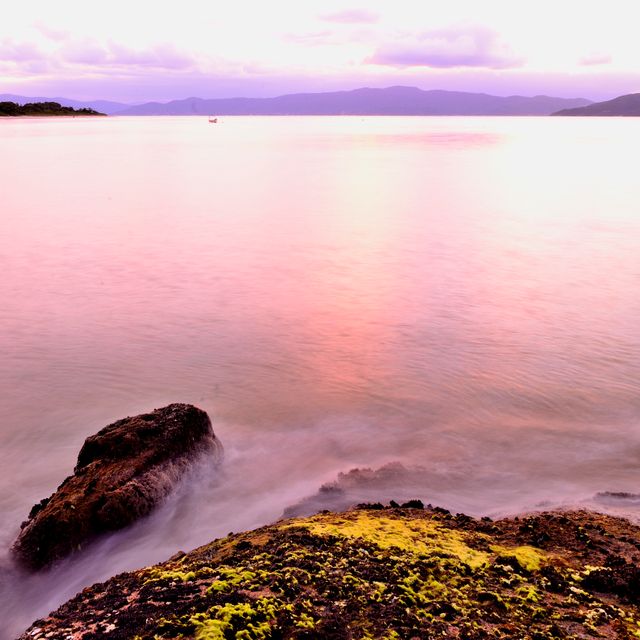 Sunset over a calm ocean with rocks and misty waters in the foreground. Mountains appear on the horizon under a colorful sky. Ideal for promoting travel destinations, relaxation retreats, or nature-themed backgrounds.