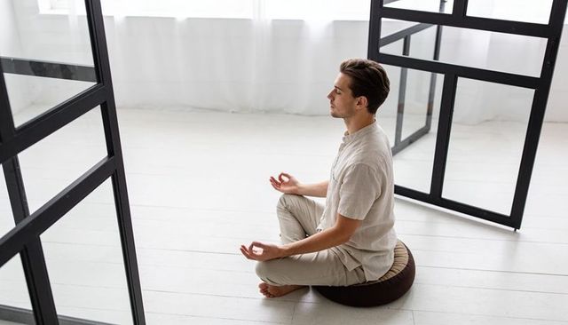 Young man meditating on round cushion in bright minimalist studio practicing mudra mindfulness