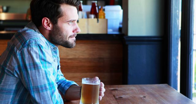 Young man in casual plaid shirt holding beer mug while gazing out window in modern pub. Ideal for themes of leisure, relaxation, contemplation, and modern lifestyle. Great for use in advertisements for pubs, bars, beer brands, or articles about urban life and personal downtime.