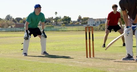 Crouching wicketkeeper wearing pads and gloves behind stumps on sunny cricket pitch