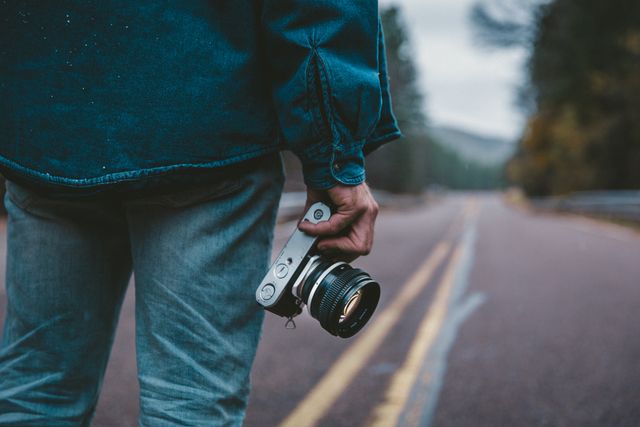Young man holding camera while standing on open road during cloudy day. Capturing adventure and spontaneous travel moments. Ideal for use in travel blogs, photography websites, lifestyle magazines, and promotional material for camera equipment or outdoor gear. Emphasizing themes of adventure, freedom, and capturing life's moments.