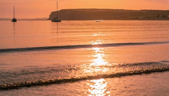 Sunset sailboats reflecting golden light across calm bay with distant cliffs and harbor