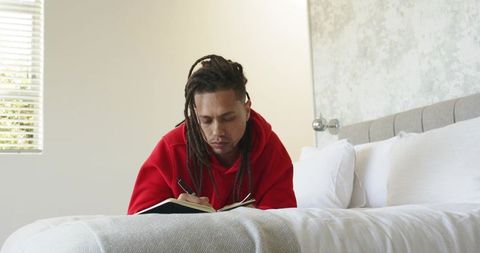 Mid 30s man writing journal on bed wearing red hoodie in minimal bright bedroom