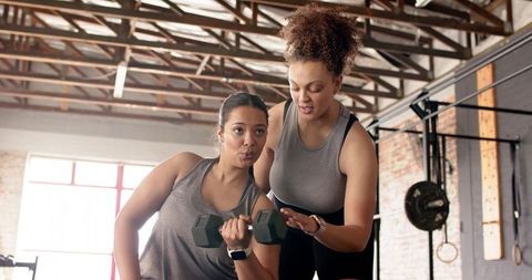Female trainer coaching trainee doing hex dumbbell curl in sunlit industrial gym