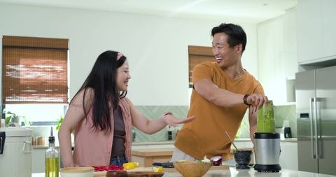 Laughing couple blending green smoothie and preparing fresh ingredients on kitchen island