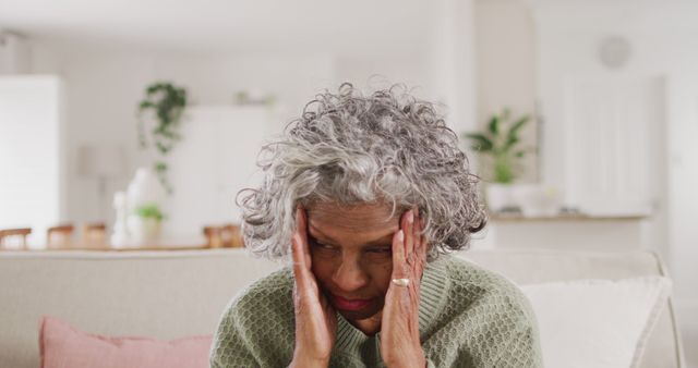 Senior woman sitting on sofa with hands on head looking stressed. Useful for illustrating topics related to mental health, loneliness, elderly care, anxiety, home life, and aging.