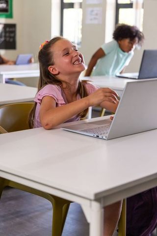 Elementary students laughing and collaborating on laptop in sunlit modern classroom