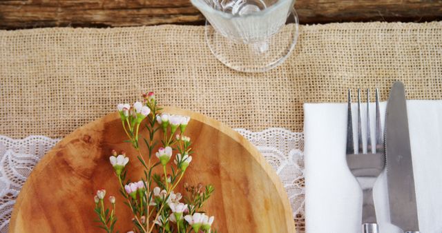 A rustic table setting features a wooden plate adorned with delicate flowers, silverware wrapped in a napkin, and an elegant glass, with copy space. The arrangement suggests a natural and simplistic dining elegance, for an outdoor event or a country-themed gathering.
