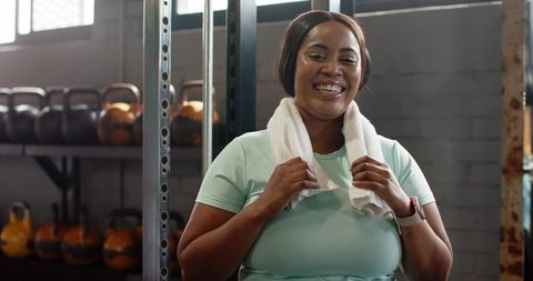 African american woman smiling after workout holding towel in gym with kettlebells in activewear