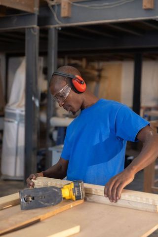 African american carpenter aligning wood plank wearing ear protection in workshop