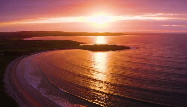 Golden sunset reflecting on curved sandy bay with headland and gentle waves