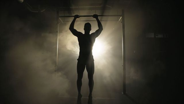 Silhouetted athlete performing pull-up in foggy industrial gym with dramatic backlight