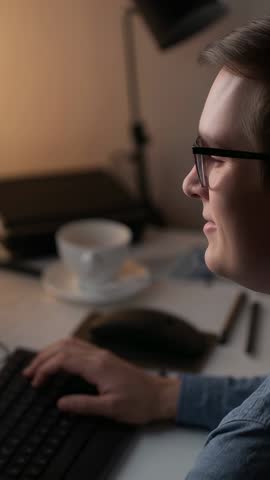 Vertical video: Man smiling while typing at home office desk under warm lamp, wearing glasses