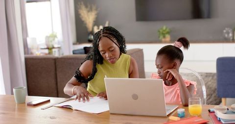 African mother tutoring daughter at home using laptop and workbook for remote learning
