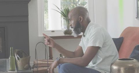 Pointing bearded man sitting on sofa, gesturing in living room with firewood and plants