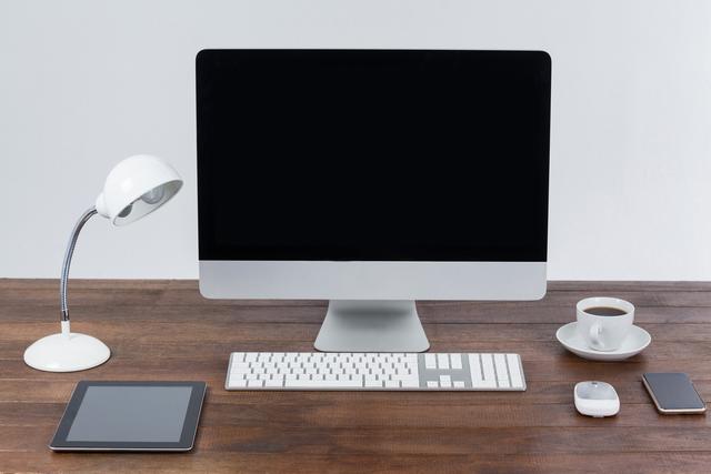 Modern office desk featuring a computer, digital tablet, mobile phone, keyboard, mouse, coffee cup, and desk lamp on a wooden surface. Ideal for illustrating concepts of technology, productivity, and modern work environments. Suitable for business presentations, office-related articles, and home office setup inspirations.