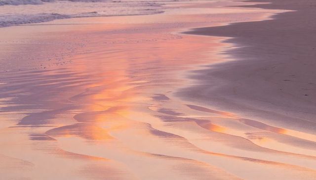 Pastel sunrise reflecting on wet sandy shoreline with gentle ripple patterns