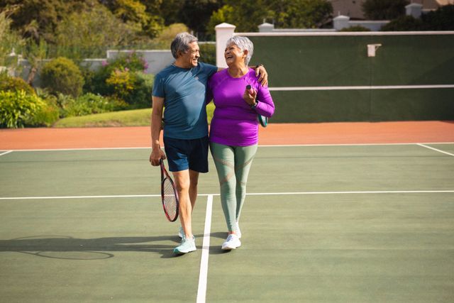 Full length of cheerful biracial senior couple with arms around and rackets walking on tennis court. sport, competition, unaltered, love, togetherness, lifestyle, retirement and active lifestyle.