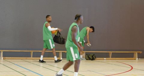Entering court with gear, diverse basketball teammates warming up, carrying bags in gym