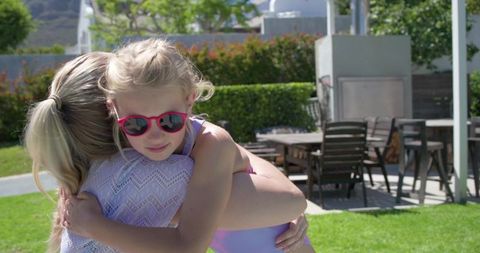 Mother hugging daughter in sunlit backyard, girl wearing red sunglasses and swimsuit