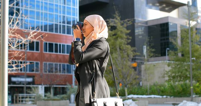This stock photo is ideal for articles, blogs, and websites focusing on cultural diversity, urban photography, female empowerment, and modern lifestyles. It features a young Muslim woman wearing a hijab and taking photographs in an urban environment with contemporary architecture.