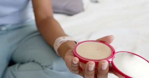 African american woman holding pink compact mirror on bed applying powder for everyday self-care