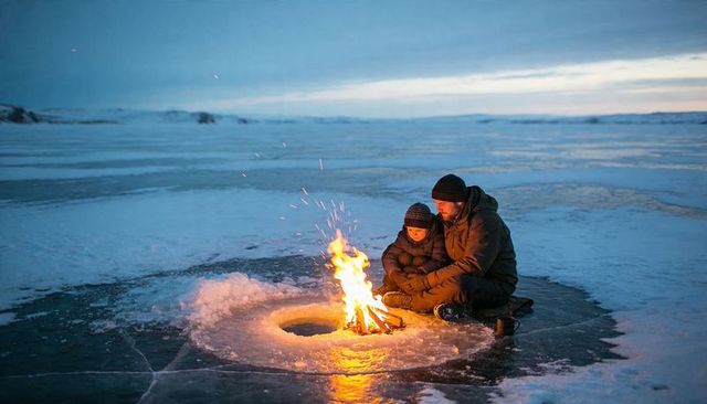 Parent and child warming by campfire over round ice hole on frozen lake at twilight