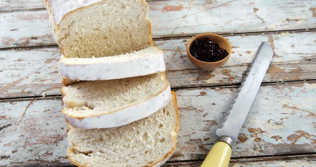 Shows freshly sliced rustic bread loaf with a serrated knife, placed on a weathered wooden table. There is a small bowl of jam near the bread. Suitable for use in blog posts, bakery advertisements, poster designs, and articles on homemade recipes and traditional baking methods.
