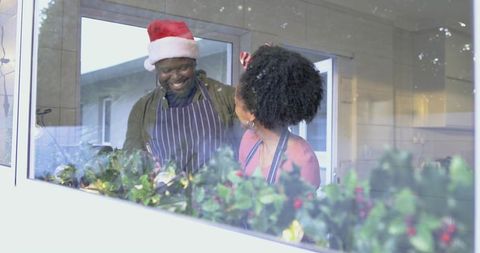 Diverse couple smiling and baking in home kitchen wearing santa hat and festive garland