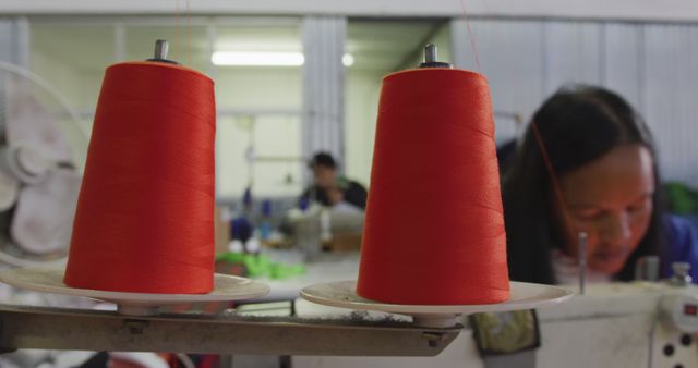Two large spools of red thread in focus in a textile factory. Workers are busy with garment production in the background, illustrating industry activity.