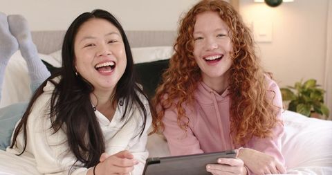 Two friends laughing on bed wearing cozy hoodies and socks while sharing tablet